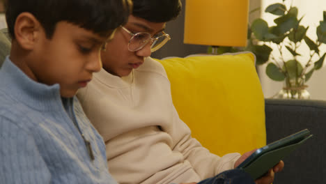 Two-Young-Boys-Sitting-On-Sofa-At-Home-Playing-Games-Or-Streaming-Onto-Digital-Tablets-13